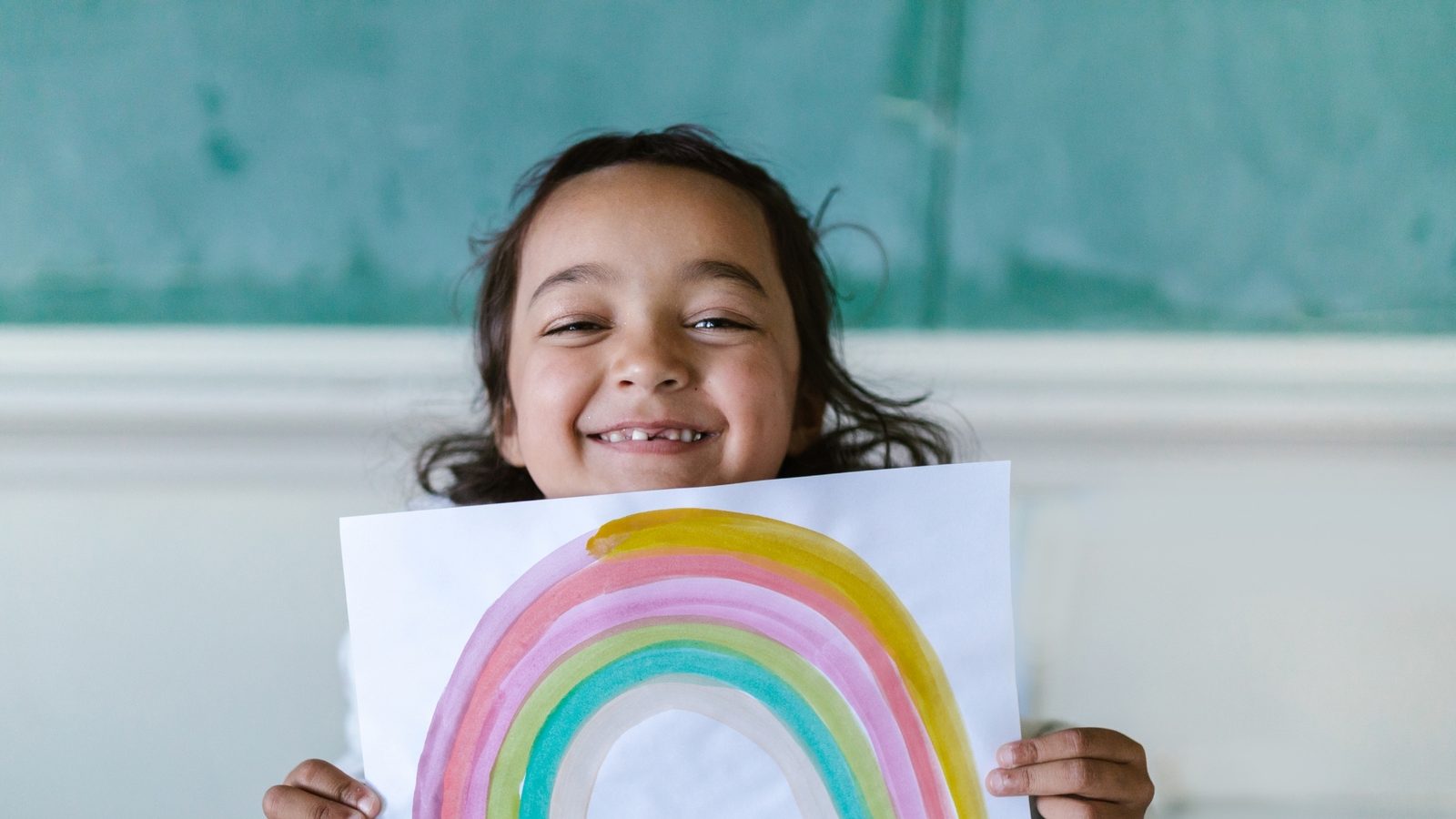 A smiling girl holding up her rainbow drawing