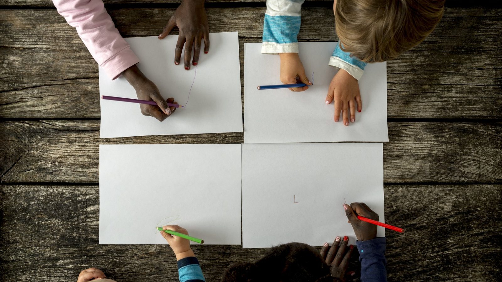 Children's hands drawing on sheets of paper on a wooden surface